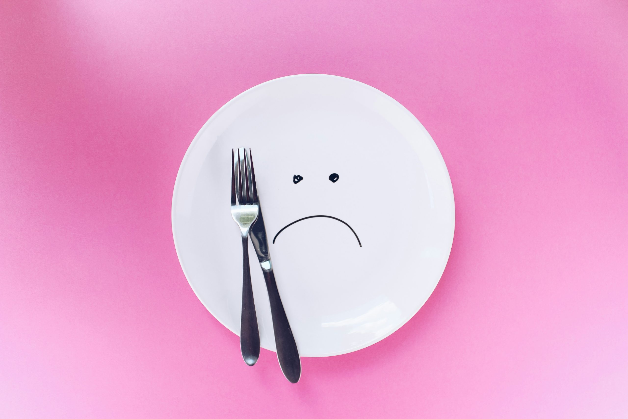 PLate with a knife and fork and a sad face drawn on it. pink background