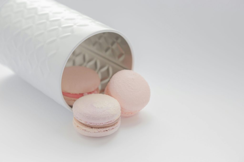 Three pastel pink macarons spilling out of a decorative white container on a soft, light background, representing tempting sugary treats and the concept of sugar cravings